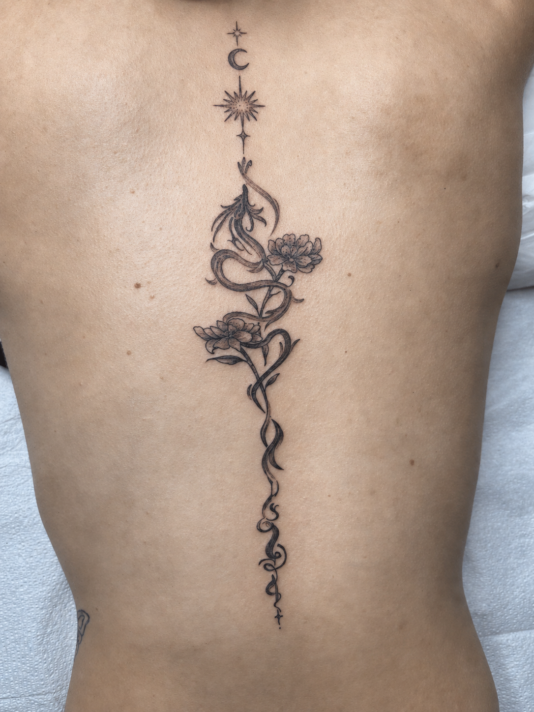 Fine line floral spine tattoo with flowers, vines, crescent moon and celestial details